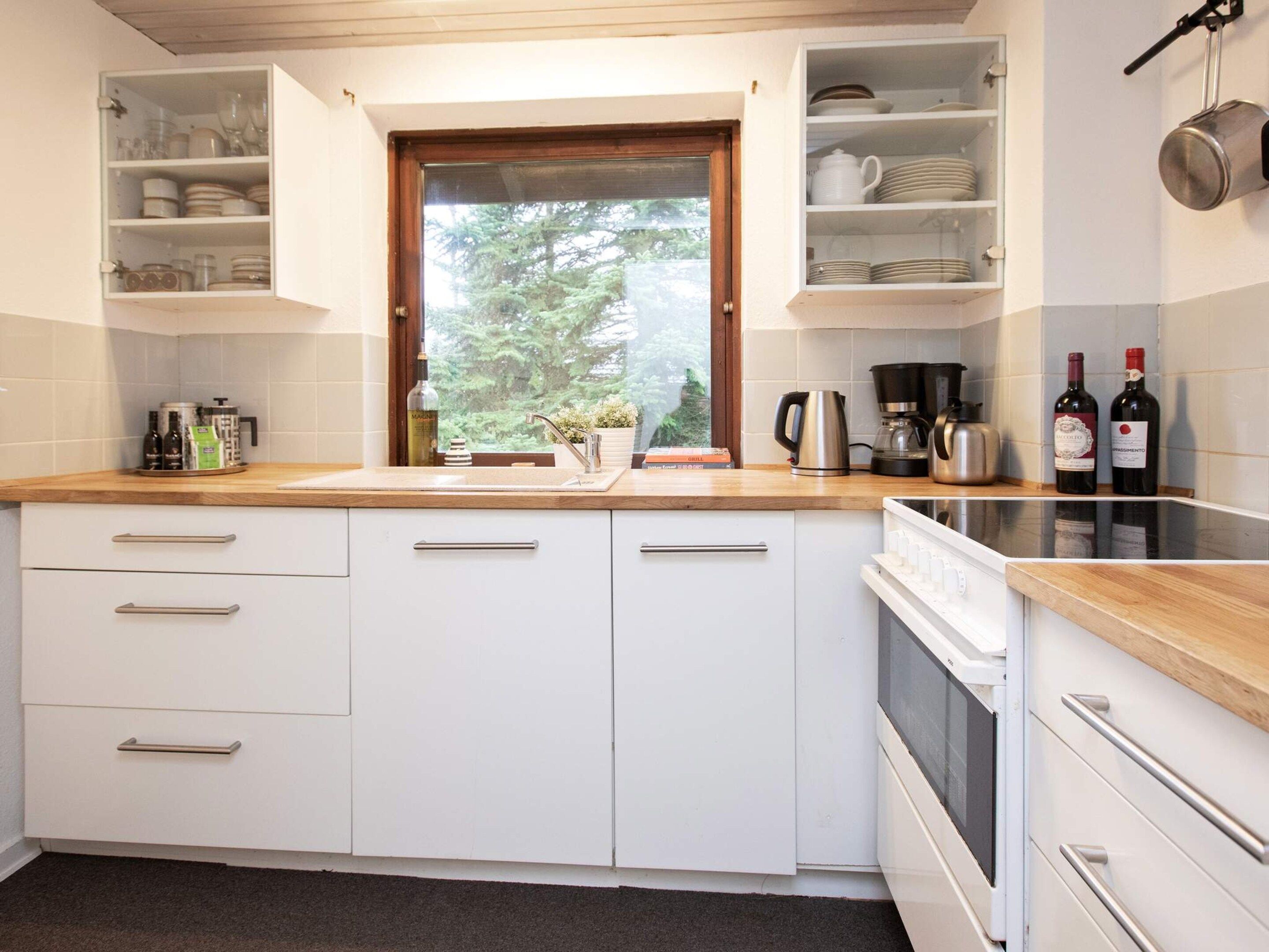 A kitchen with white cabinets, a wooden countertop, a window, and a view of trees.