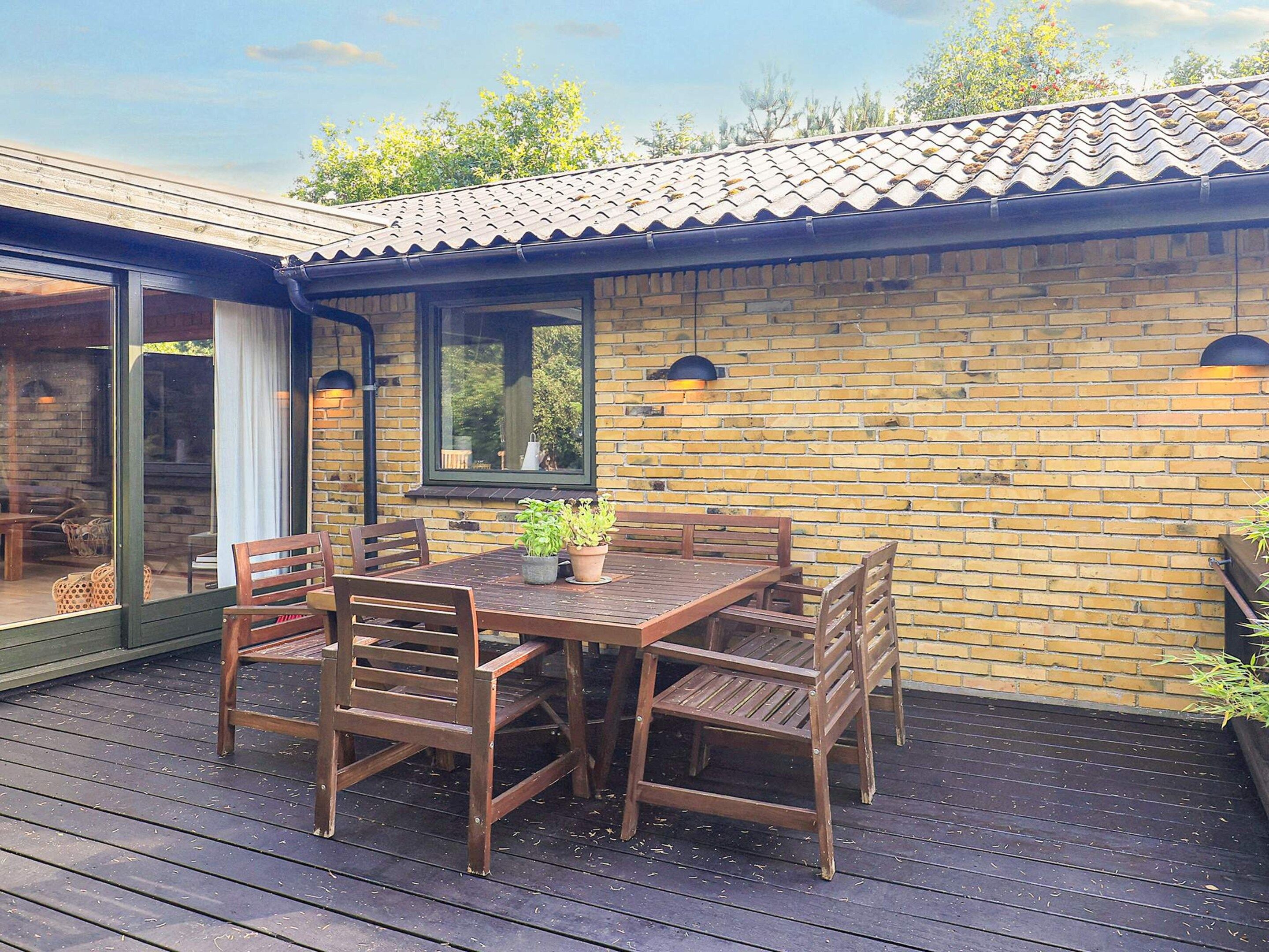 Une terrasse en bois avec une table et des chaises, un mur en briques et une fenêtre donnant sur un paysage verdoyant.