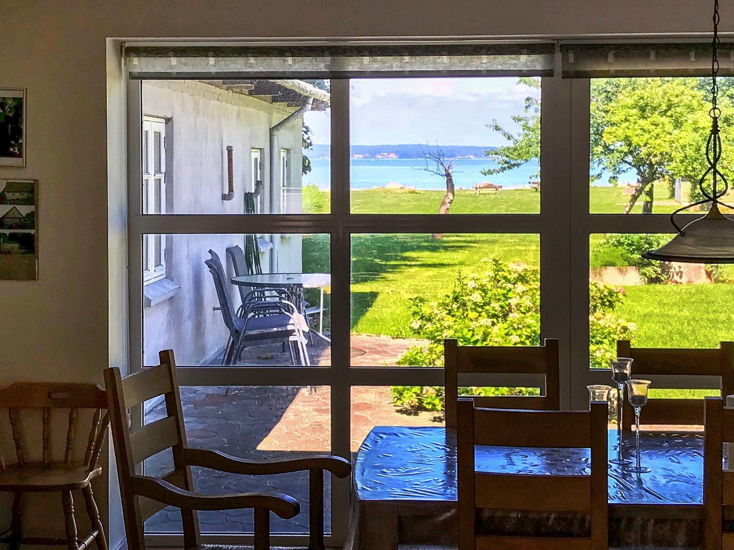Ein Zimmer mit großem Fenster, das auf eine Wiese und ein Gebäude blickt. Im Raum befinden sich Stühle und ein Tisch.