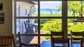 Ein Zimmer mit großem Fenster, das auf eine Wiese und ein Gebäude blickt. Im Raum befinden sich Stühle und ein Tisch.
