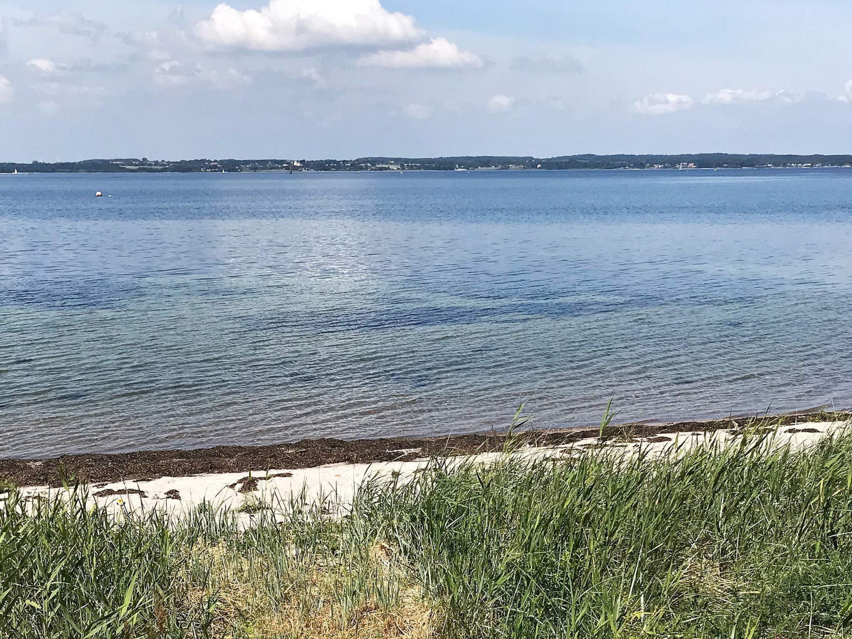 Roligt vand med en sandstrand i forgrunden og en fjerntliggende kystlinje med bygninger.