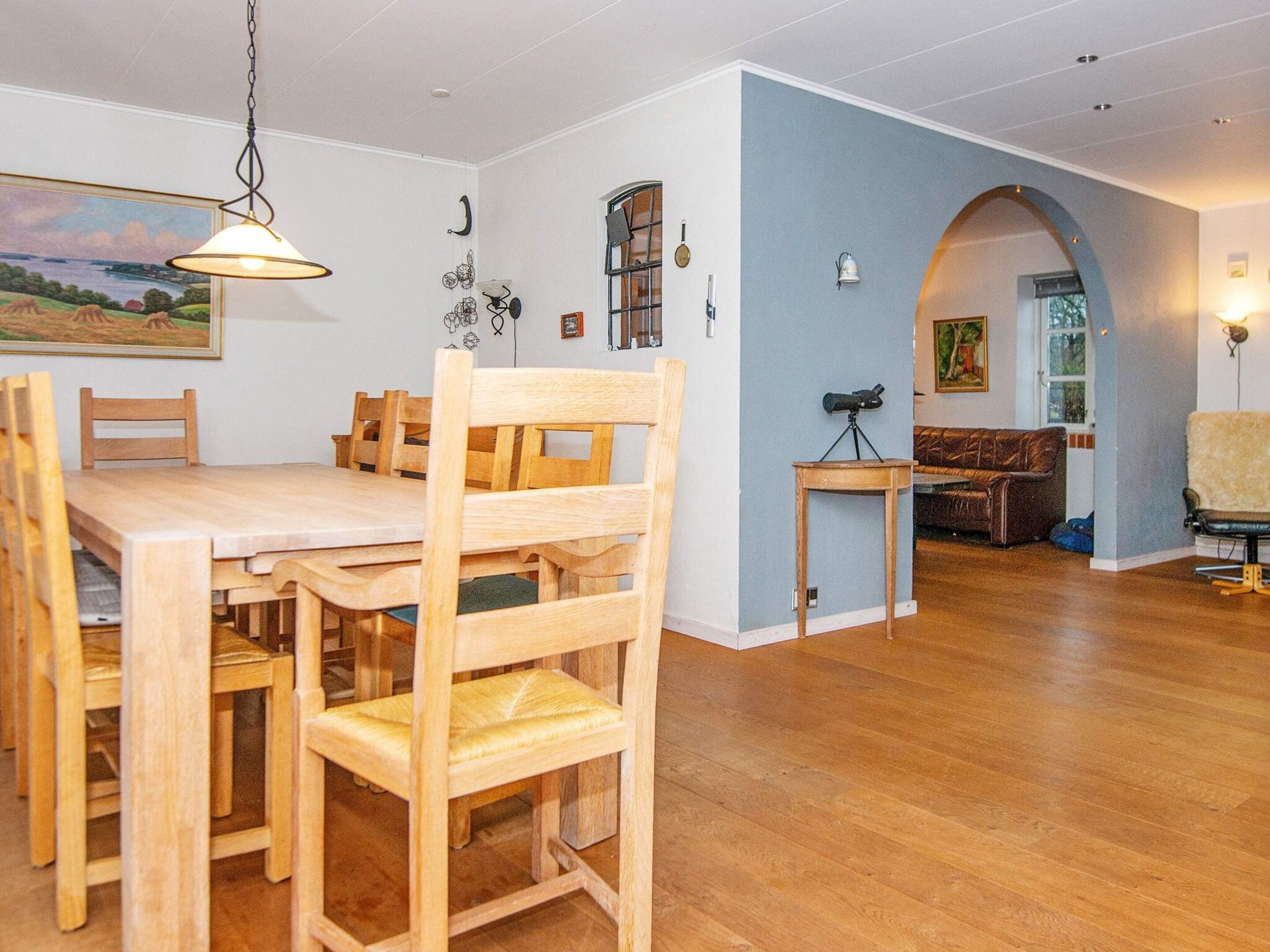A dining area with a wooden table and chairs, a hanging light fixture, and a painting on the wall.
