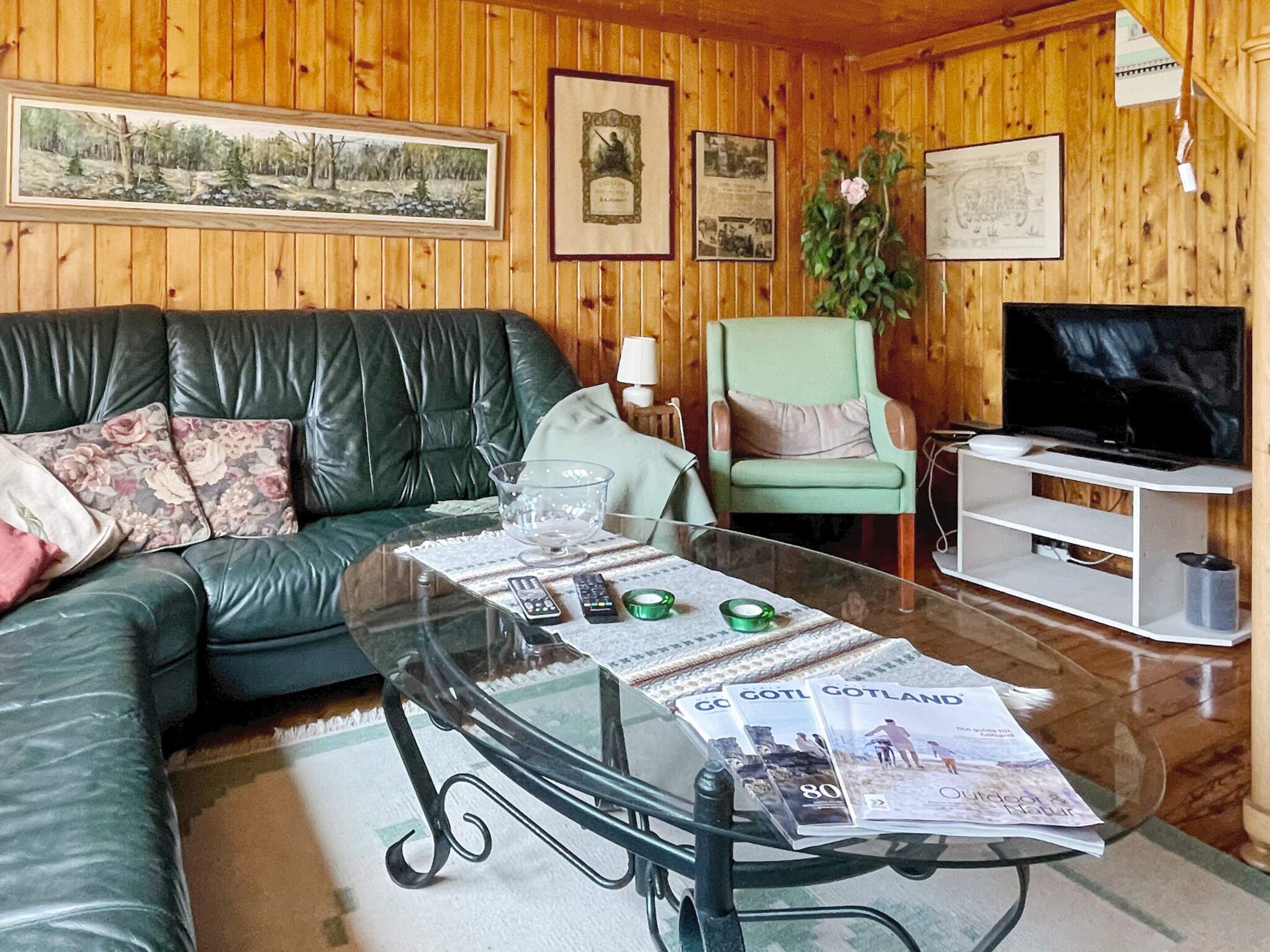 Een woonkamer met een leren bank, een glazen salontafel met tijdschriften, een groene fauteuil, een televisie en houten wanden.