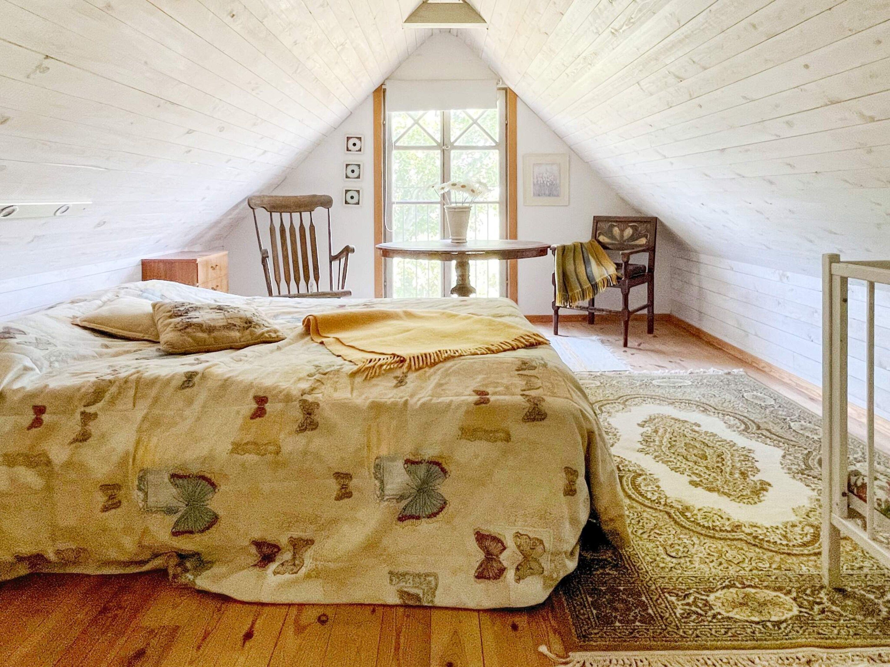 Een knus zolderkamer met een bed, een stoel, een klein tafeltje en een raam.