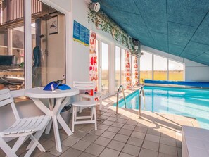 An indoor swimming pool area with a tiled floor, a pool with a blue edge, and a covered seating area with white chairs and tables.