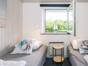 A room with two single beds, a small table, a window with a view of greenery, and a wall-mounted light.