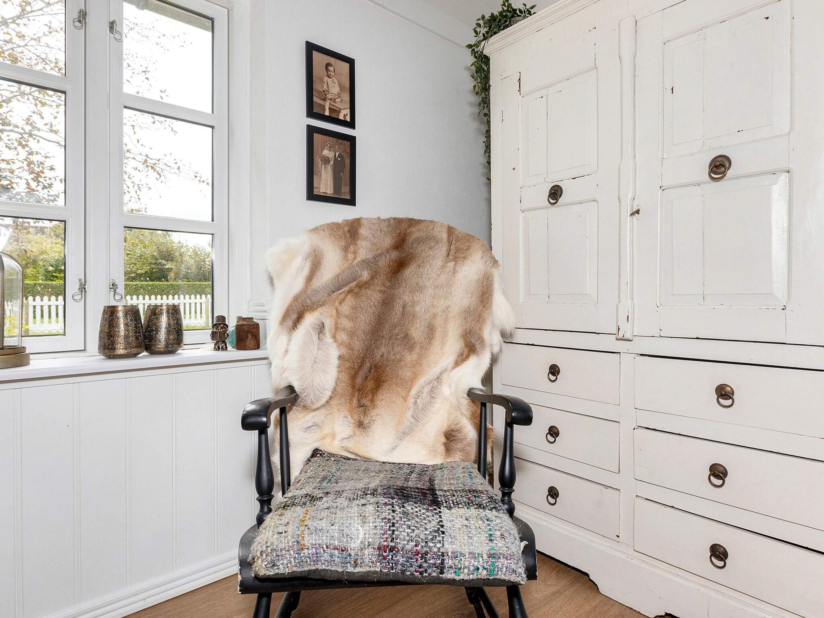 Una silla con una manta de piel, un armario blanco con cajones y dos fotografías enmarcadas en la pared.