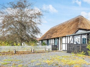 Un bâtiment traditionnel à toit de chaume, avec une clôture en bois peinte en blanc, une allée de gravier et un grand arbre en arrière-plan.