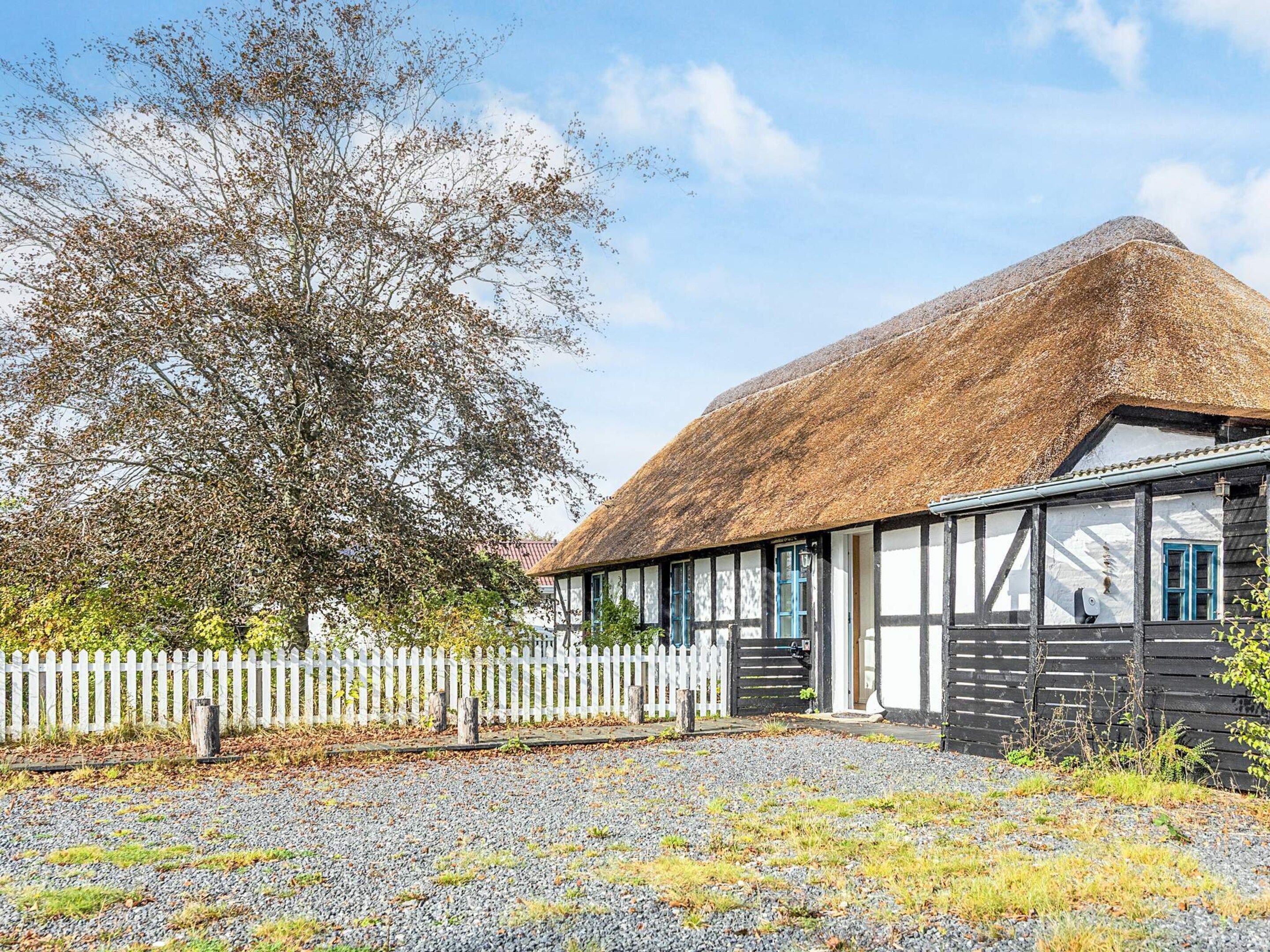 A traditional thatched-roof building with a white picket fence, gravel driveway, and a large tree in the background.