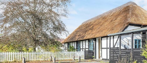 A traditional thatched-roof building with a white picket fence, gravel driveway, and a large tree in the background.