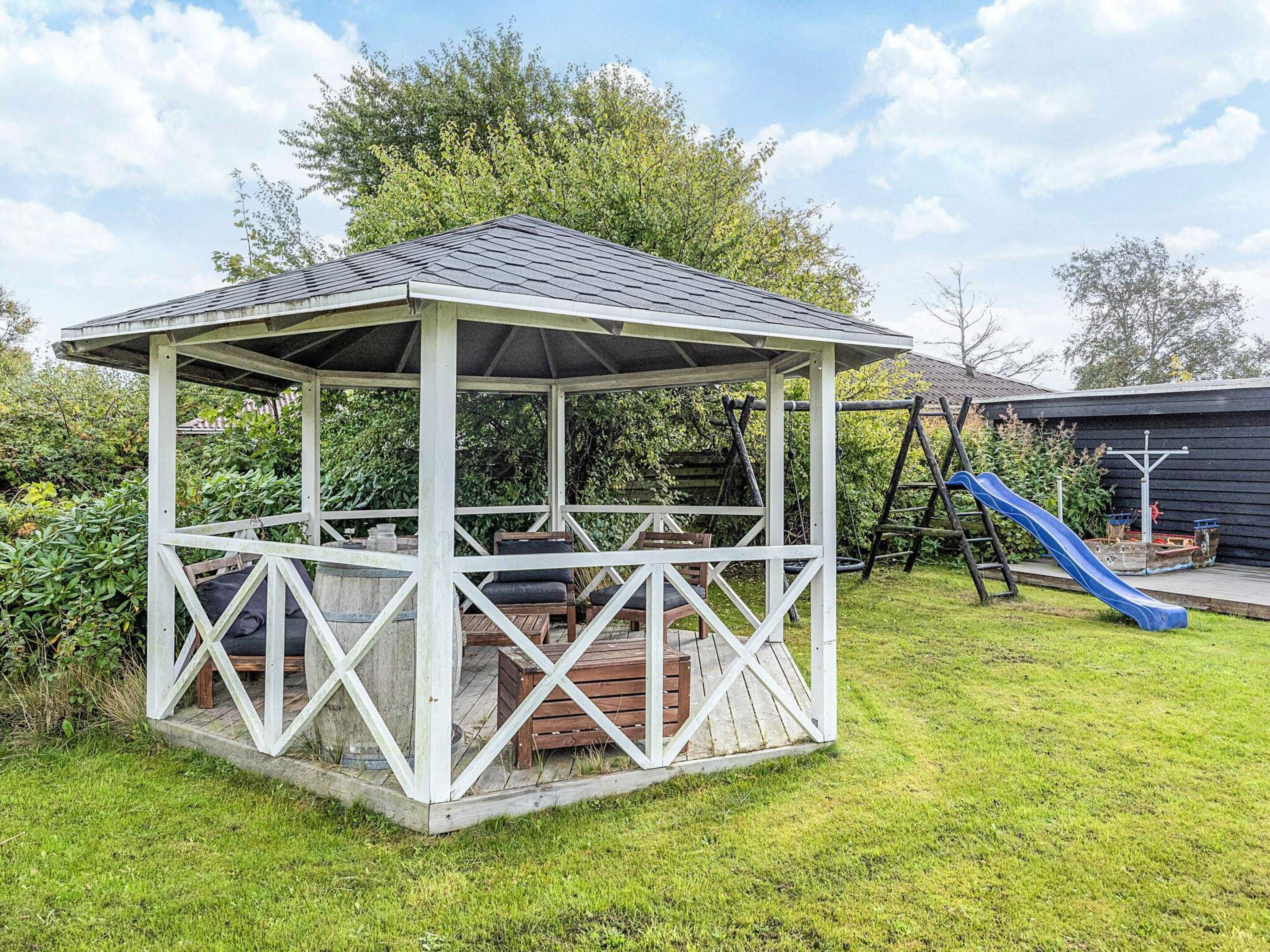 A gazebo with a roof and a wooden deck, surrounded by a grassy yard and trees.