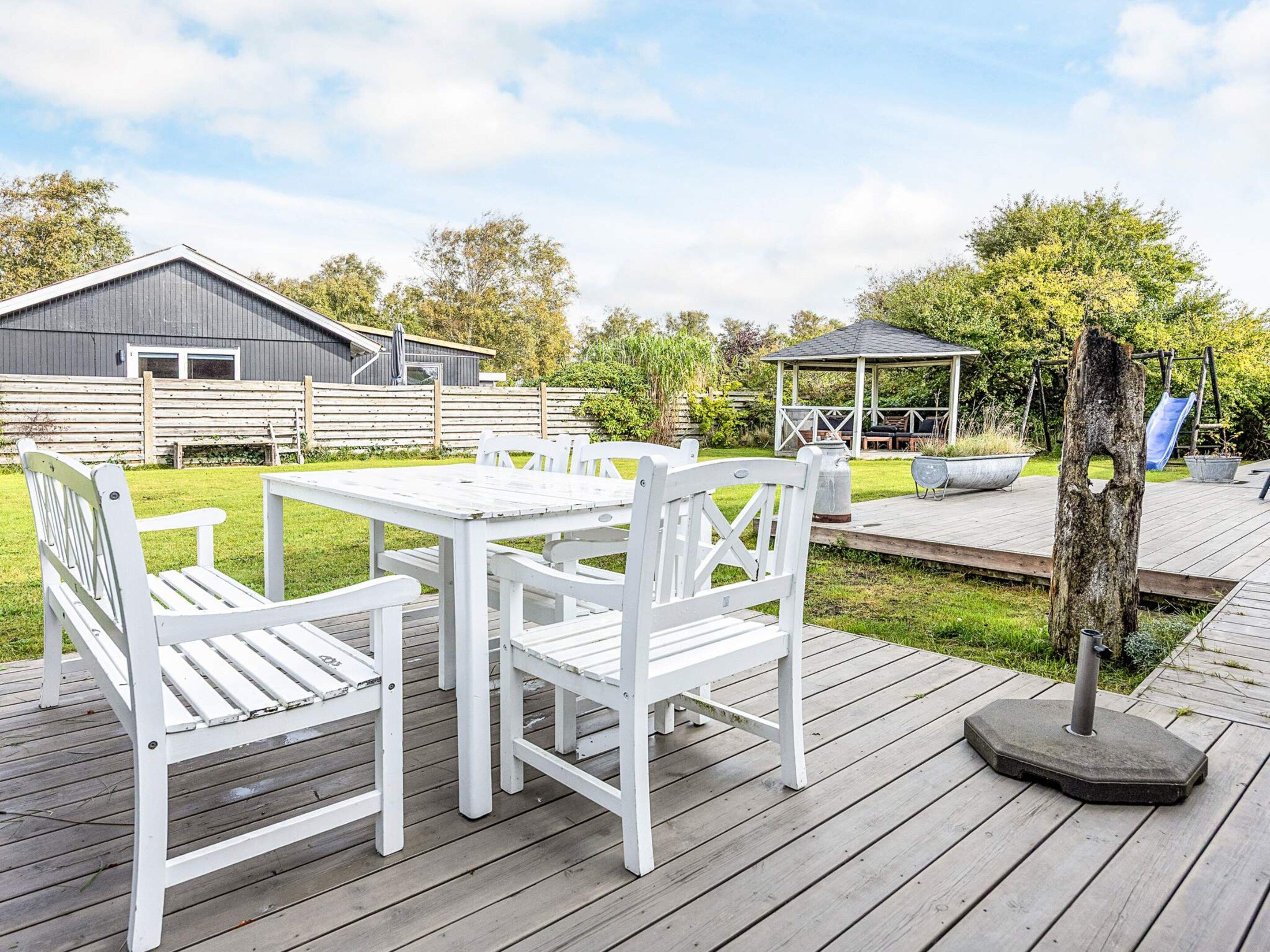 En træterrasse med et hvidt bord og stole, en pergola og en rutschebane i baggrunden.