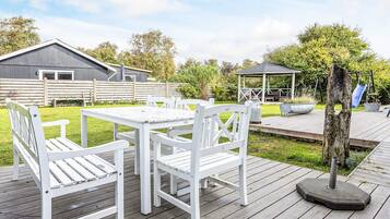En træterrasse med et hvidt bord og stole, en pergola og en rutschebane i baggrunden.