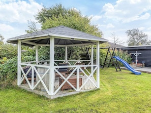 A gazebo with a sloped roof, wooden flooring, and a swing set in the background.