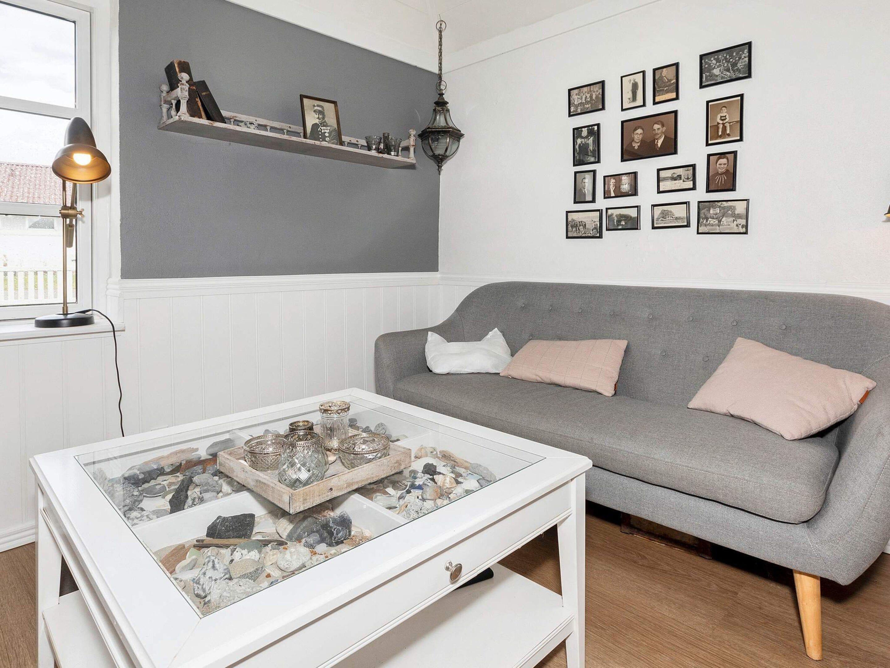 En stue med en grå sofa, et glasbord med sten, en vægmonteret hylde med billeder og en hængende lampe.