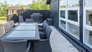 An outdoor patio with a dining table and wicker chairs, a barbecue grill, and a large window overlooking a garden.