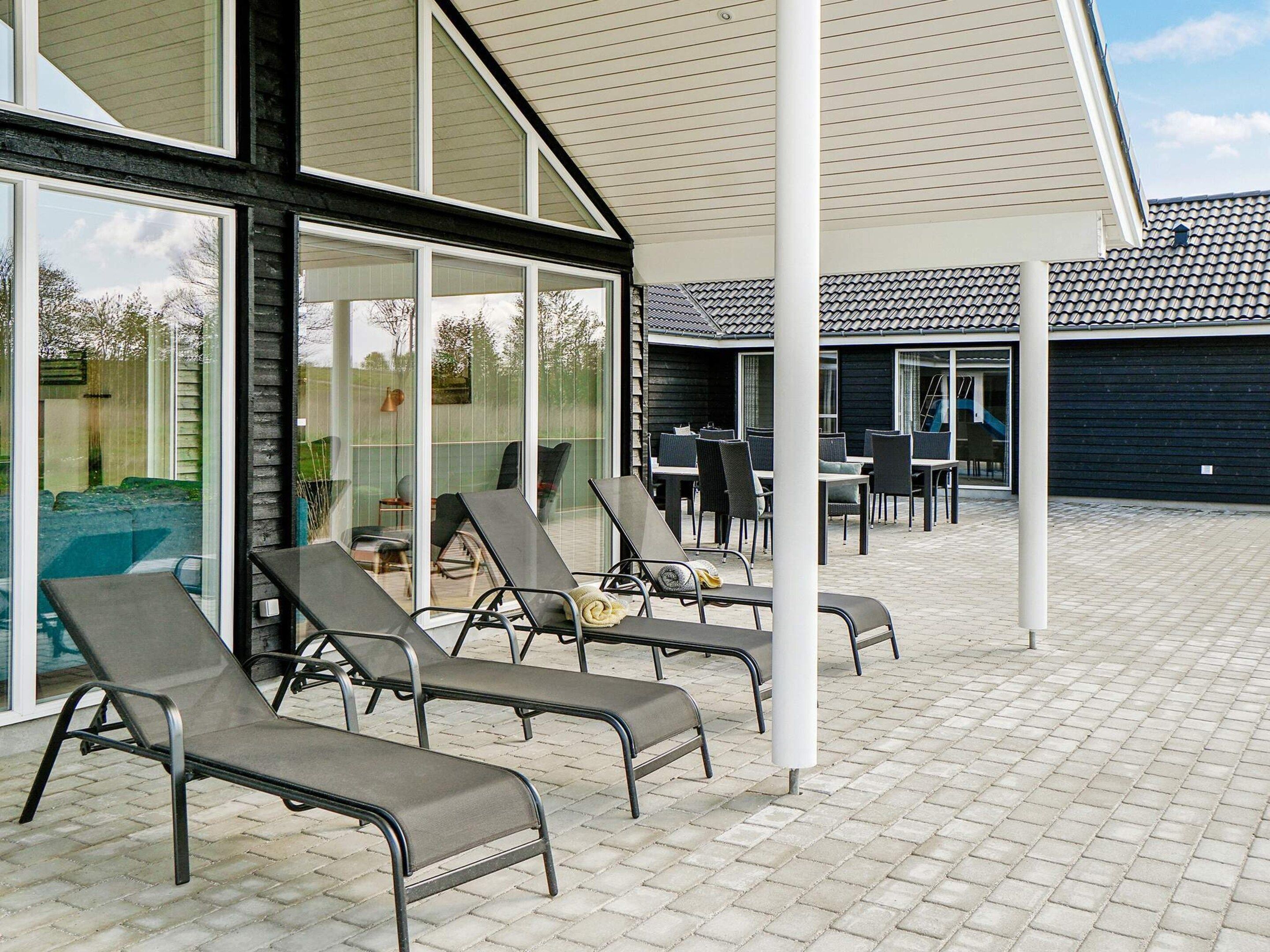A covered outdoor seating area with lounge chairs and tables, a glass door entrance, and a tiled roof.