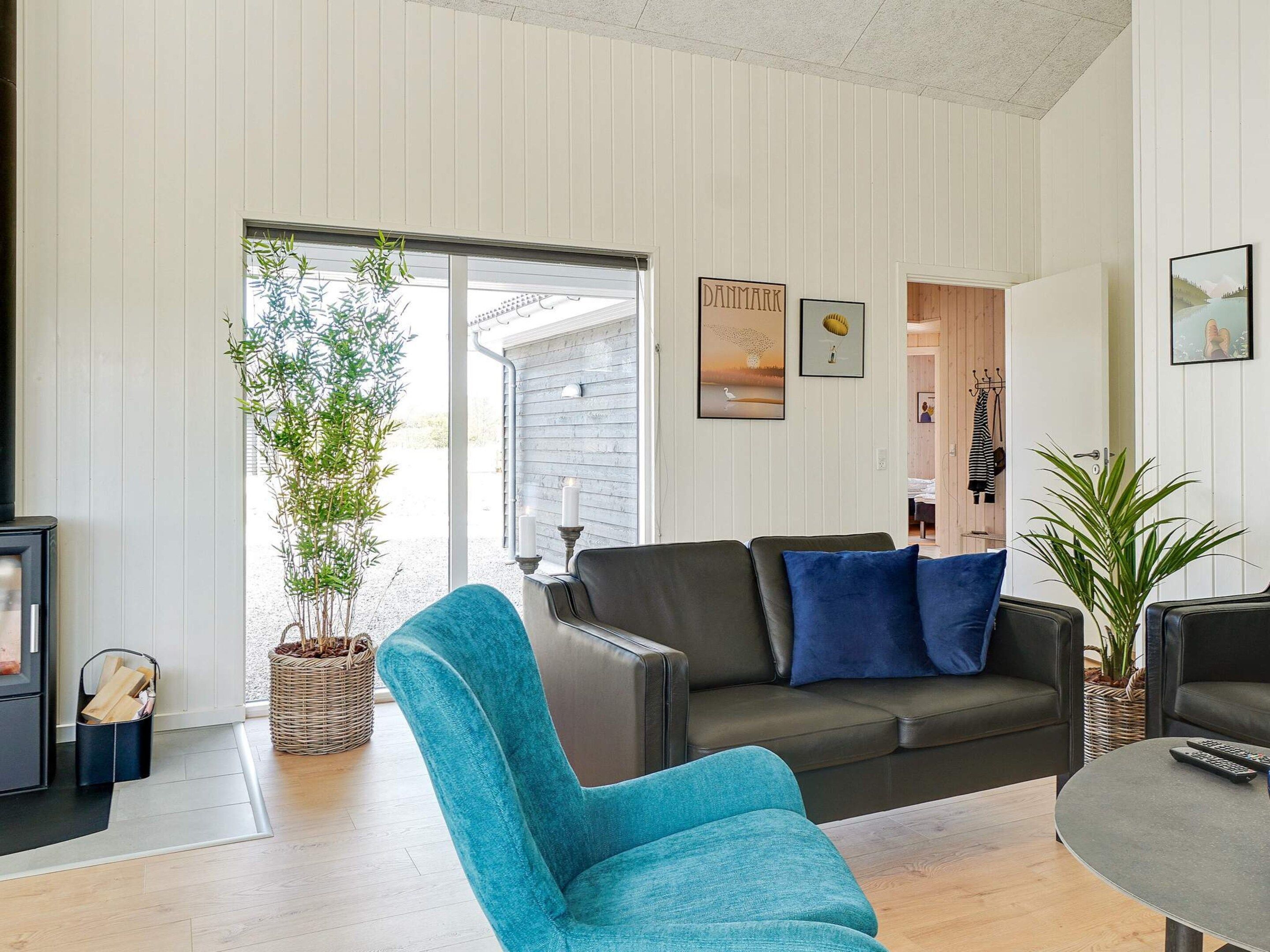A modern living room with a sofa, a blue armchair, and a coffee table. There are framed pictures on the wall, a plant, and a view into another room.