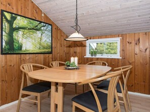 A wooden-paneled room with a round table, six chairs, a hanging light fixture, a framed picture of a forest, and a window with a view of trees. - 6 Person Holiday Home in Romo (Rømø)
