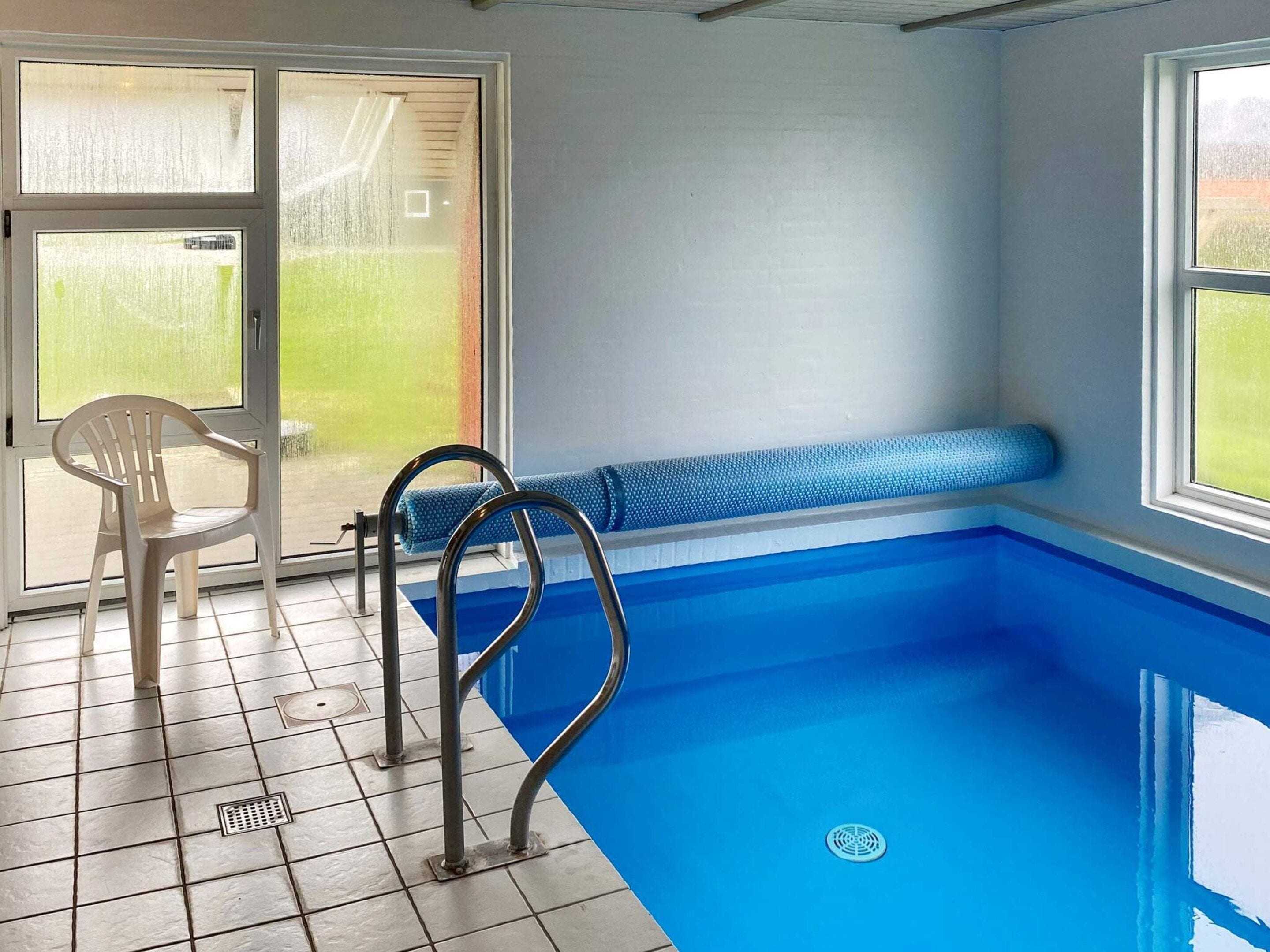 En swimmingpool med en bænk og en stol, flisegulv og et vindue med udsigt til grønne områder.
