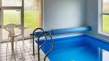 En swimmingpool med en bænk og en stol, flisegulv og et vindue med udsigt til grønne områder.