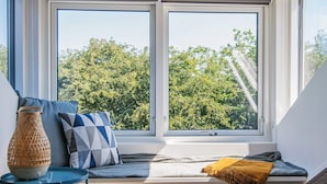 A room with a large window, a blue sofa with cushions, a woven basket, and a small table. - Holiday Home in Juelsminde (Horsens)