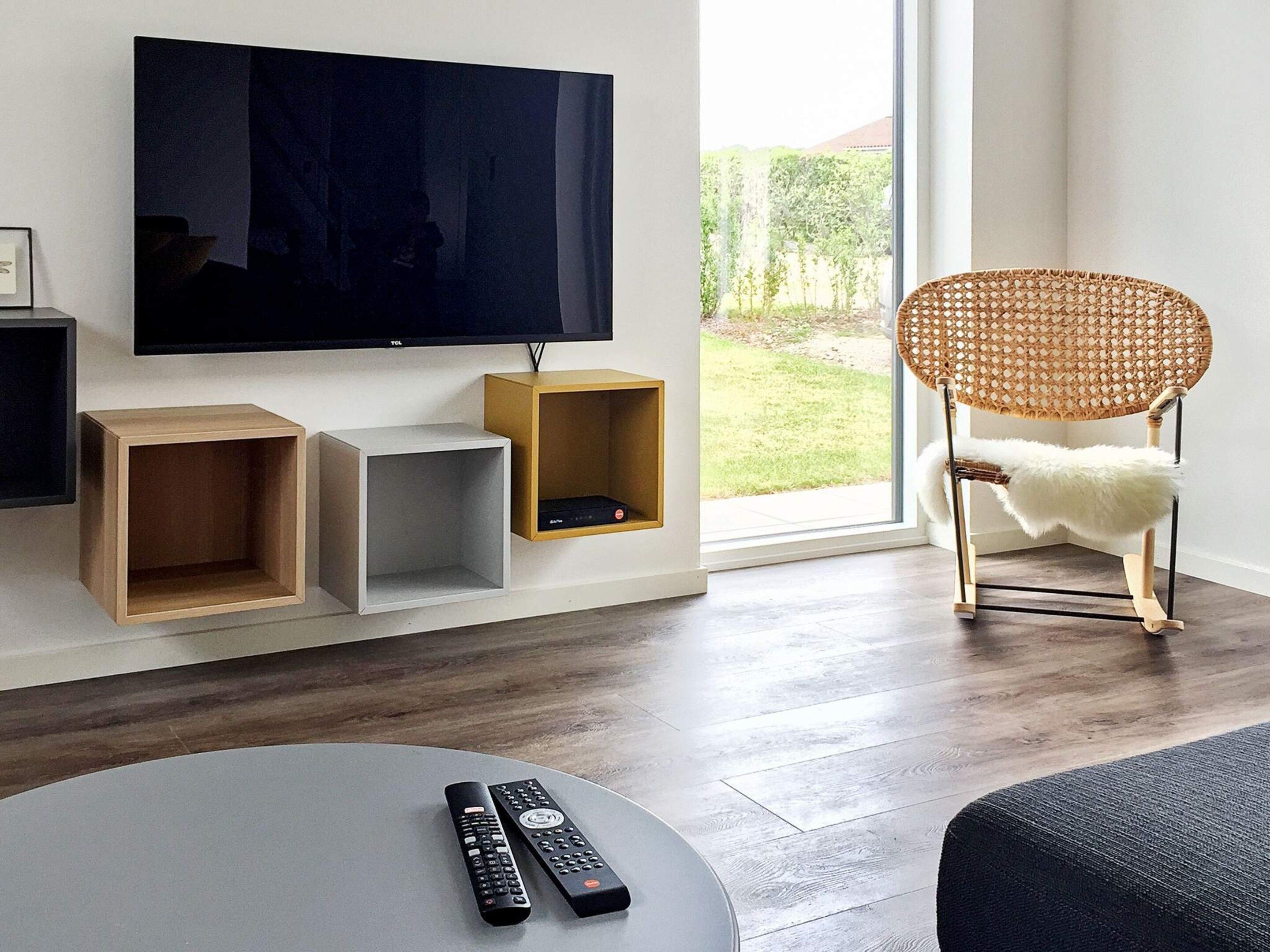 A modern living room with a flat-screen TV mounted on the wall, wooden and white storage units, a wicker chair with a white fur throw, and a round coffee table with two remote controls.