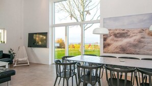 Un espace de repas moderne, doté d’une grande table et de chaises, d’un téléviseur fixé au mur et offrant une vue sur un champ verdoyant grâce à de grandes fenêtres.