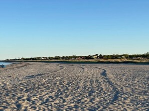 A sandy beach with footprints leading to the water, a clear sky, and a distant line of houses.
