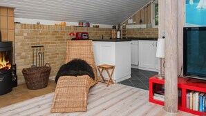A cozy living room with a fireplace, a wicker sofa, a television, and a kitchen area with a brick wall and wooden accents. - 6 Person Holiday Home in Ebeltoft (Ebeltoft)