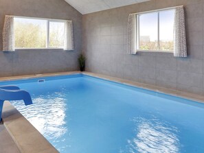 An indoor swimming pool with clear blue water, two windows with curtains, and a potted plant. - Holiday Home in Idestrup (Idestrup)