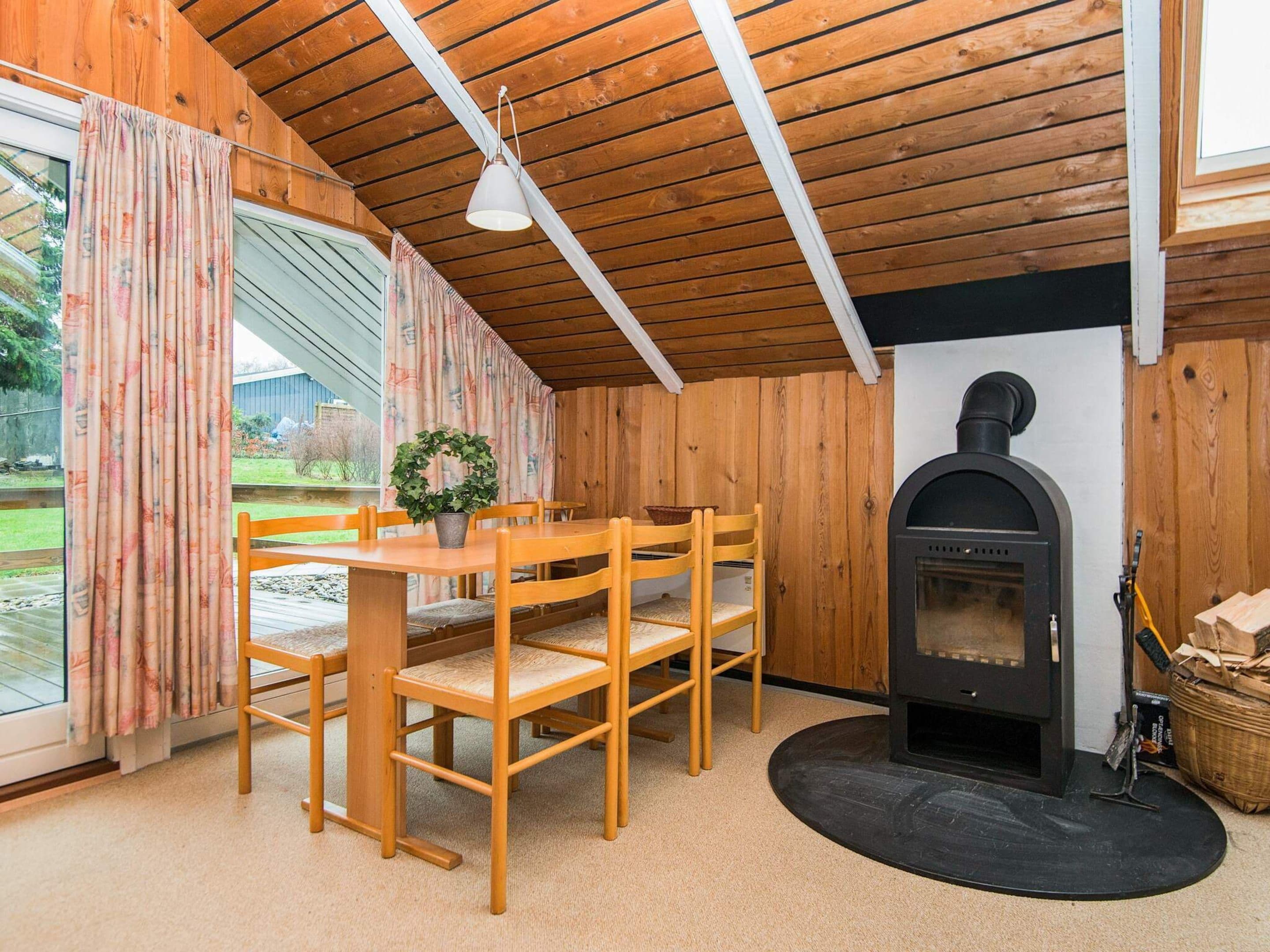 A cozy room with a wooden ceiling, a fireplace, a dining table with chairs, and a large window with curtains.