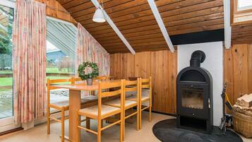A cozy room with a wooden ceiling, a fireplace, a dining table with chairs, and a large window with curtains.