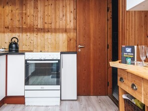 A compact kitchen with a stove, oven, and a kettle on the countertop. A wooden door and a small wooden table with a book and glasses are visible. - 6 Person Holiday Home in Blavand (Blavand)