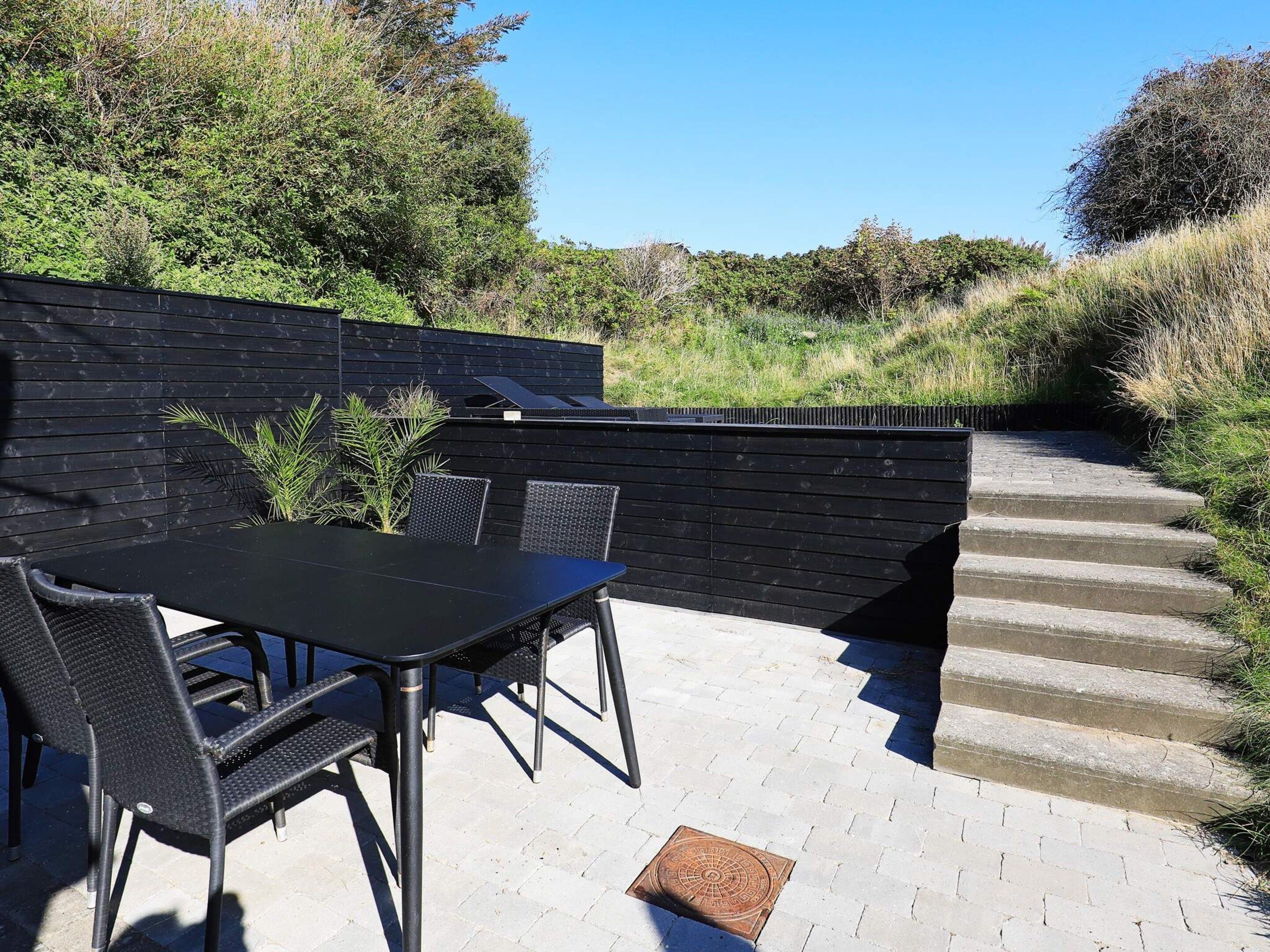 En udendørs terrasse med et bord og stole, en sort væg og et sæt trapper.