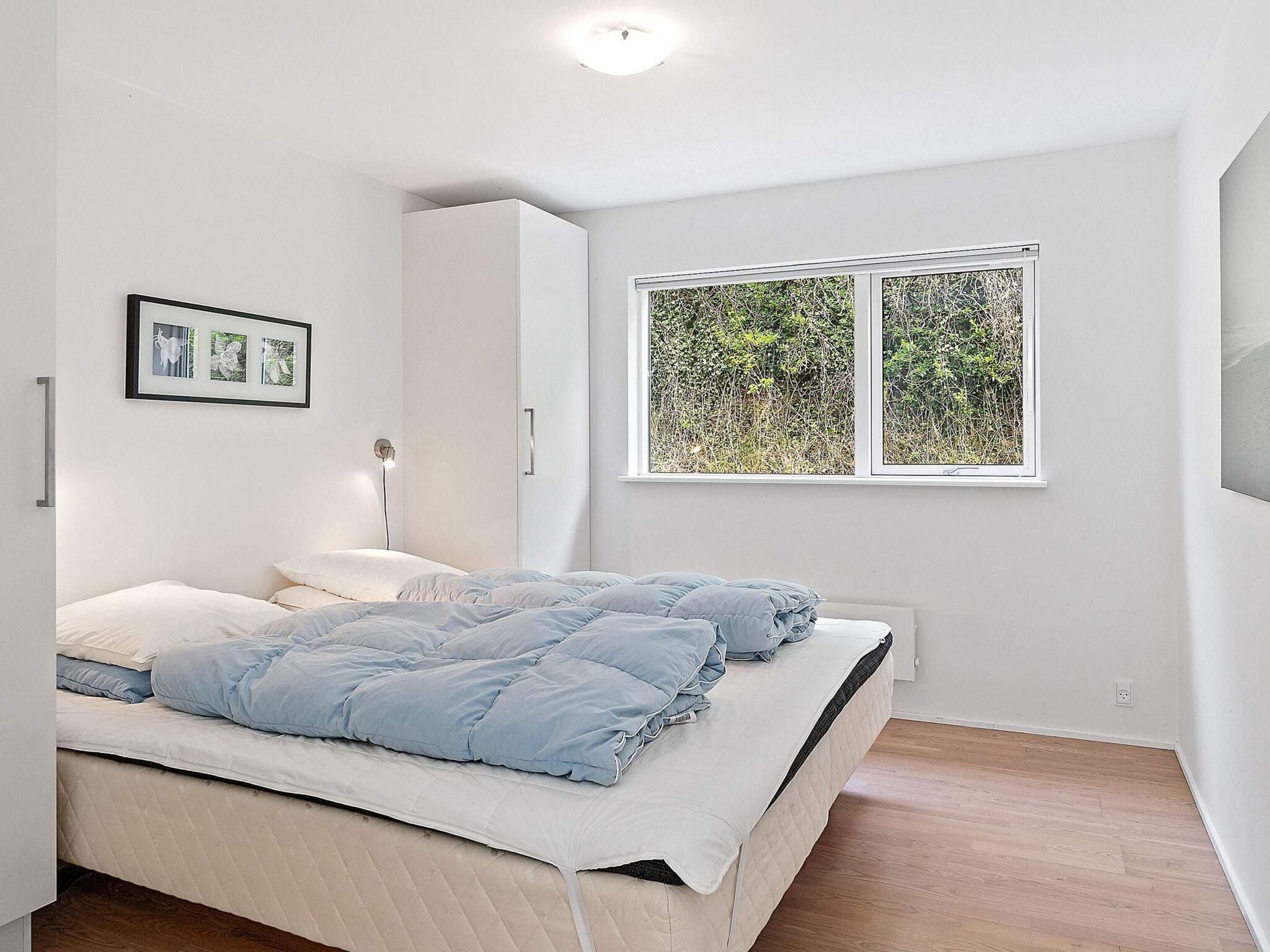 A bedroom with a bed, a bedside lamp, a wardrobe, and a window with a view of greenery.
