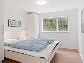 A bedroom with a bed, a bedside lamp, a wardrobe, and a window with a view of greenery. - Holiday Home in Blokhus (Blokhus)