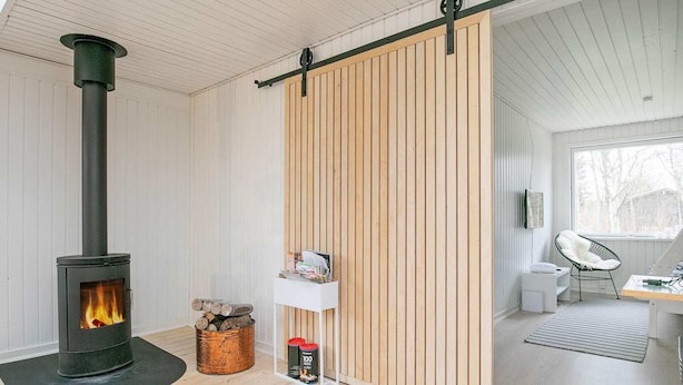 A modern living room with a wood-burning stove, a sliding wooden door, a white desk with a chair, and a large window. - 7 Person Holiday Home in Frederikshavn (Frederikshavn)