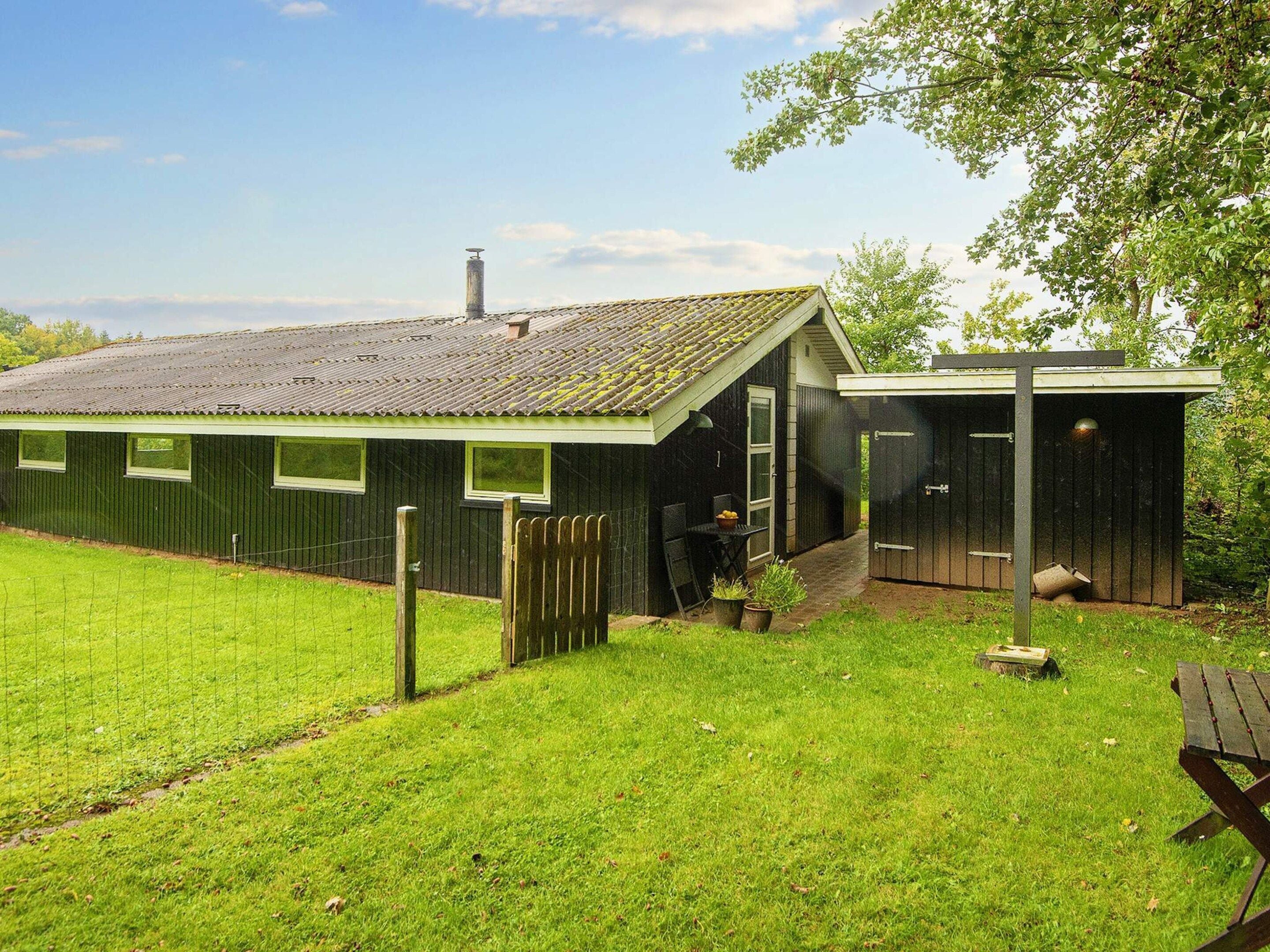 A green metal building with a sloped roof, a fenced yard, and a small garden area with a bench.