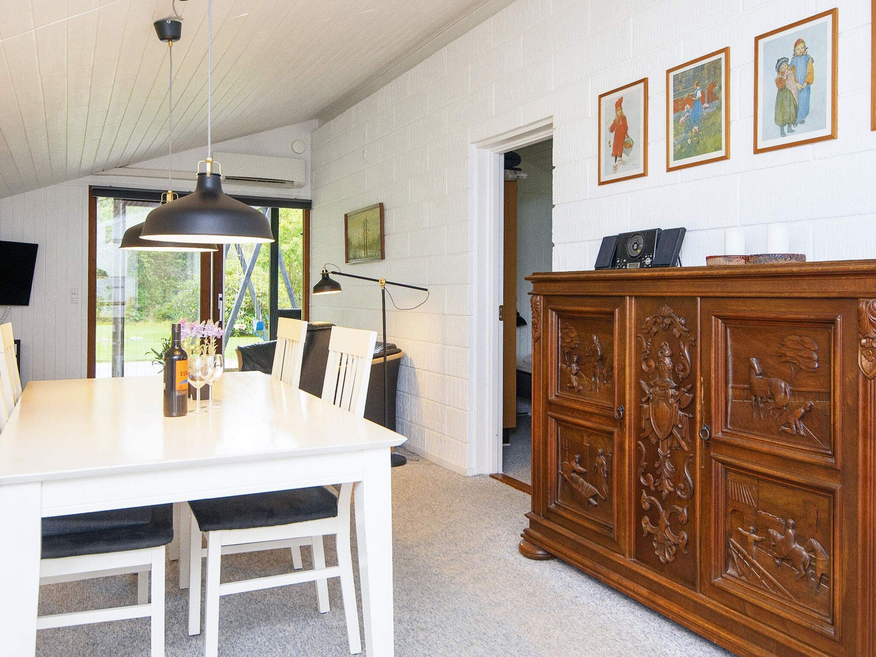 Un coin repas avec une table blanche, des chaises et un luminaire suspendu. On y trouve également un meuble en bois orné de sculptures et un téléviseur fixé au mur.