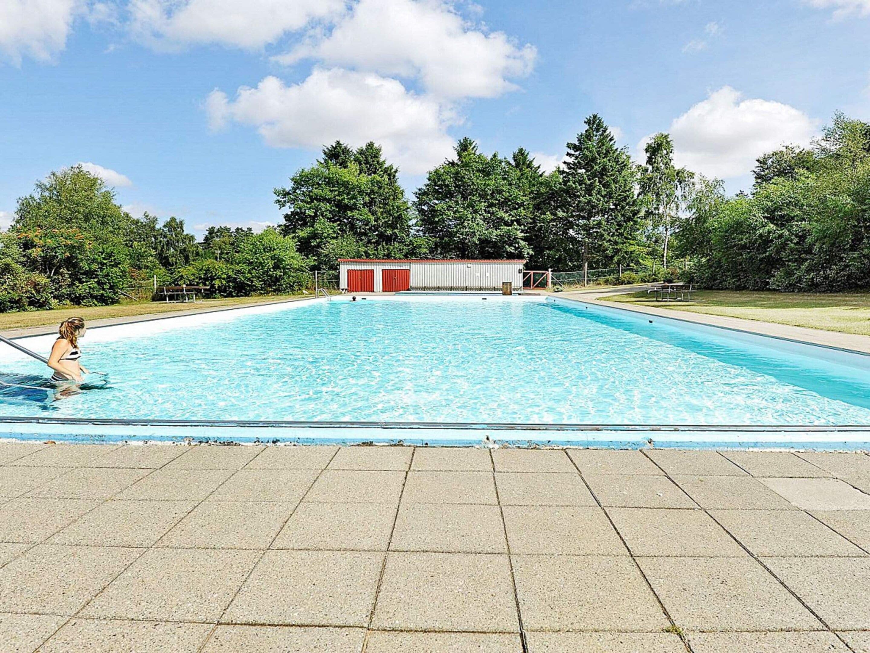 A swimming pool with a tiled deck area, surrounded by trees and a small building with red doors.