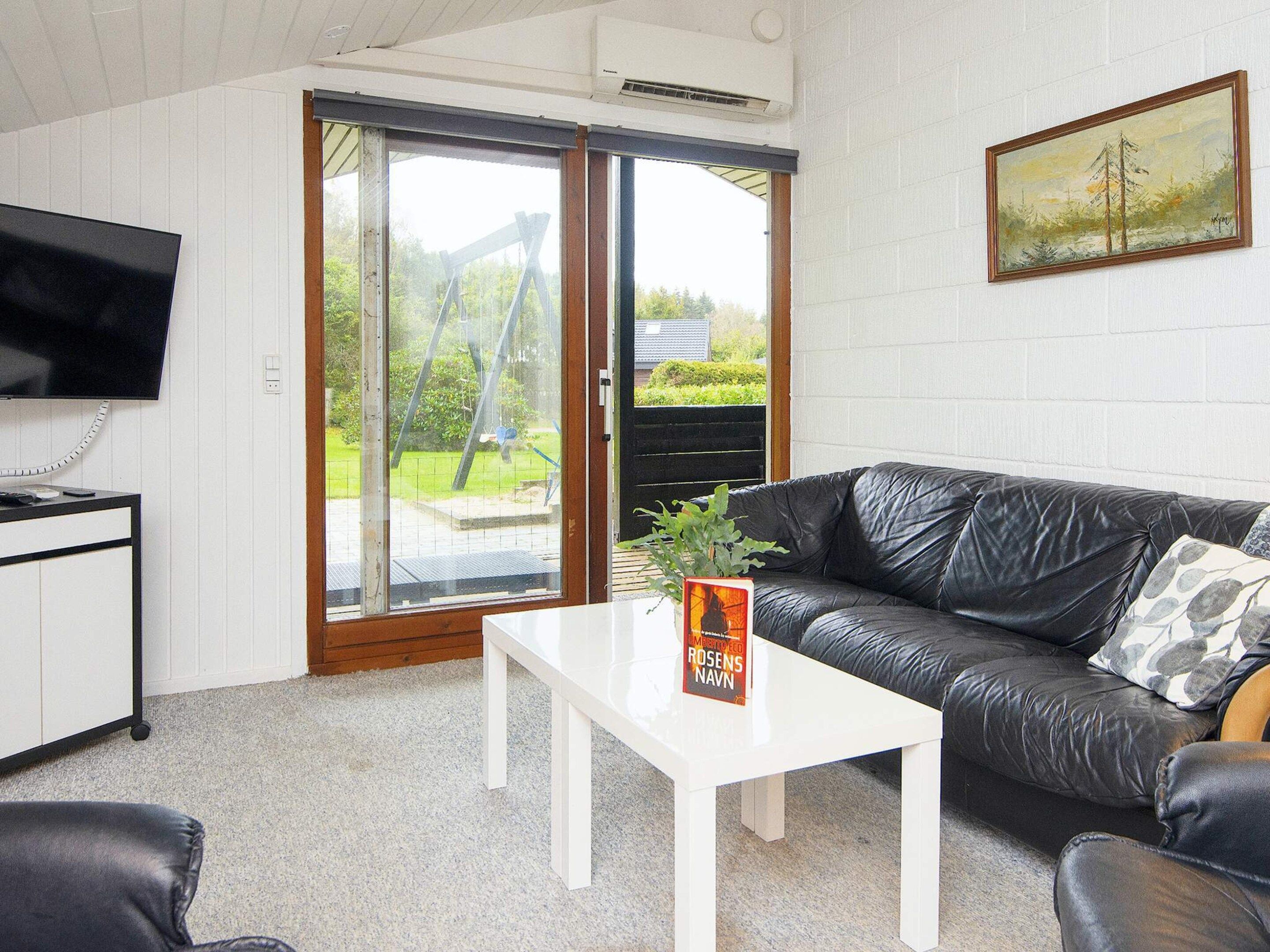 A living room with a black leather sofa, a white coffee table, a flat-screen TV mounted on the wall, and a sliding glass door leading to an outdoor area.