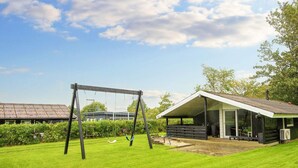 A grassy backyard with a swing set, a covered patio area, and a building with a sloped roof. - 6 Person Holiday Home in Farvang (Farvang)