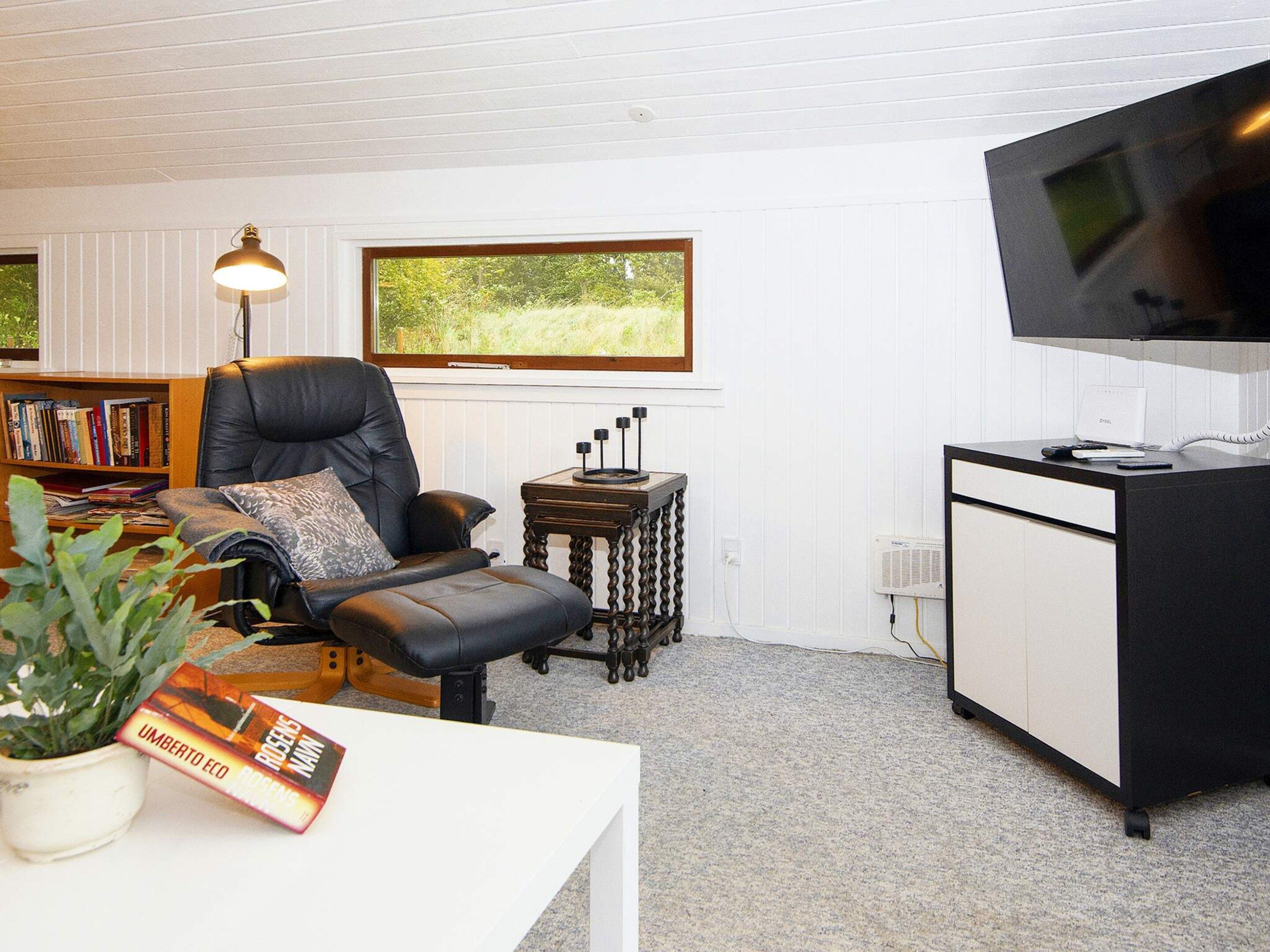 A room with a black leather chair, a white table with a plant, a bookshelf, a desk with a flat-screen TV, and a window with a view of greenery.