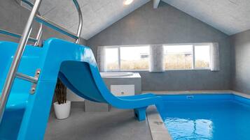 An indoor swimming pool with a blue slide, a window with curtains, and a ceiling with recessed lighting.