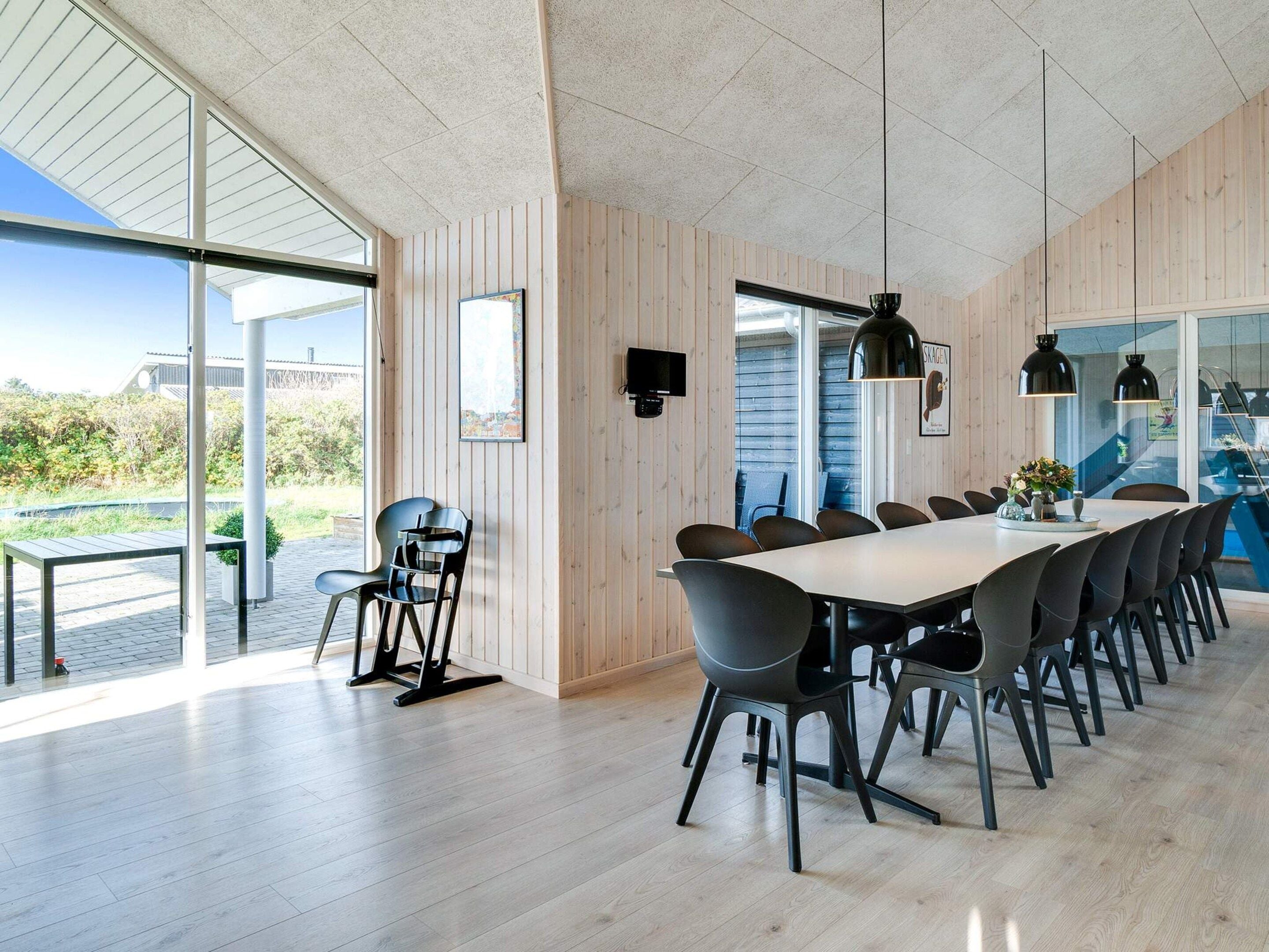 A modern dining area with a long table and chairs, large windows, and a view of the outdoors.