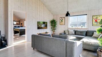 A modern living room with a grey sofa, a coffee table, and a TV on the wall. There is a window with a view of a house outside.