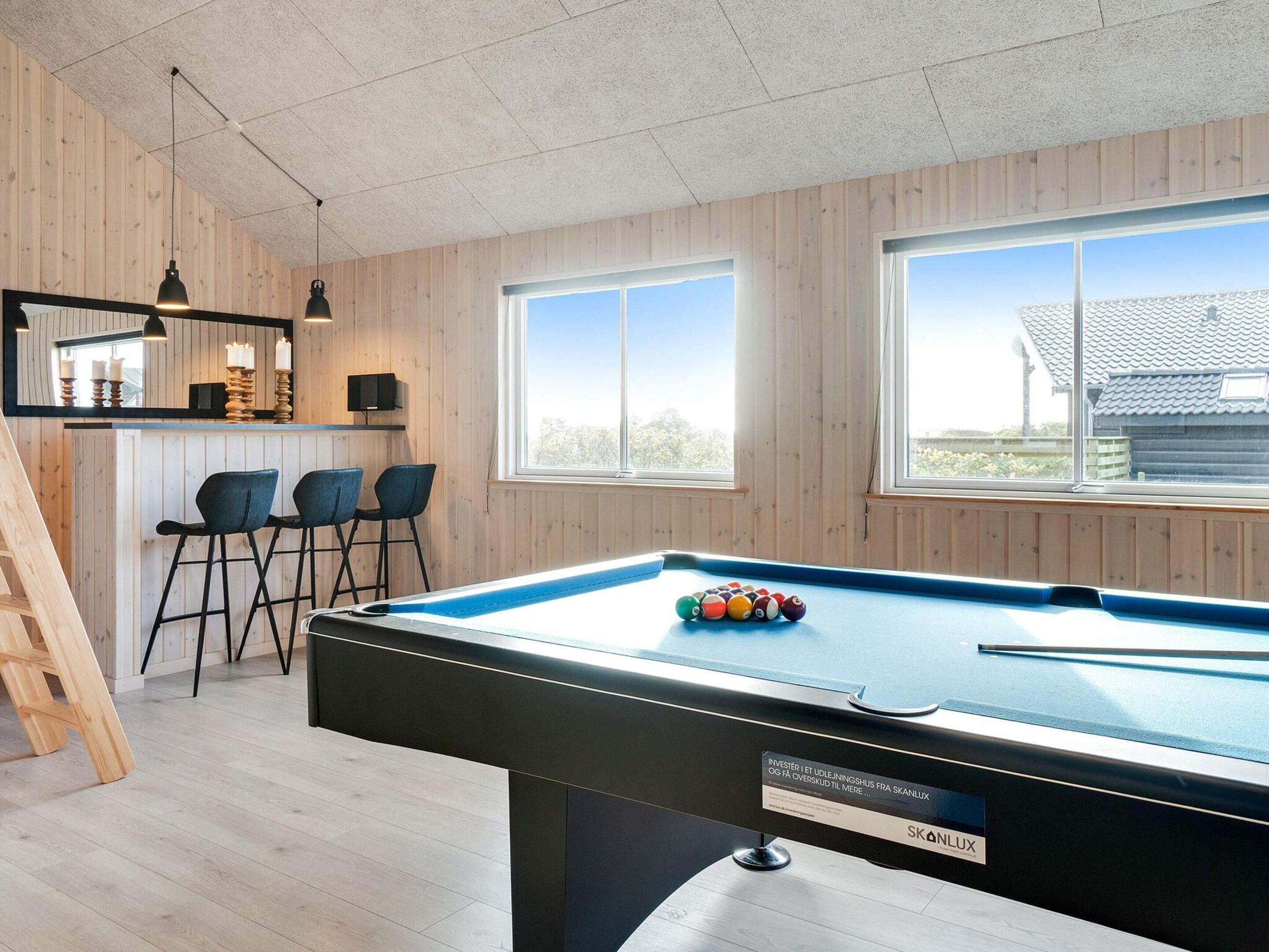 A pool table with a pool cue and balls, a bar area with stools, and a staircase leading to an upper level.
