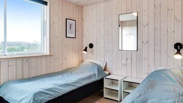 En säng med ett mönstrat överkast, ett sängbord, en spegel som hänger på väggen och ett fönster med utsikt över grönområden.