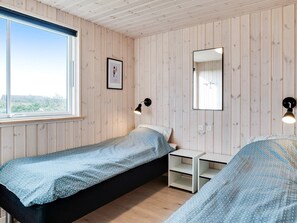A single bed with a patterned bedspread, a bedside table, a wall-mounted mirror, and a window with a view of greenery. - Holiday Home in RingkÃ¸bing (Ringkobing)